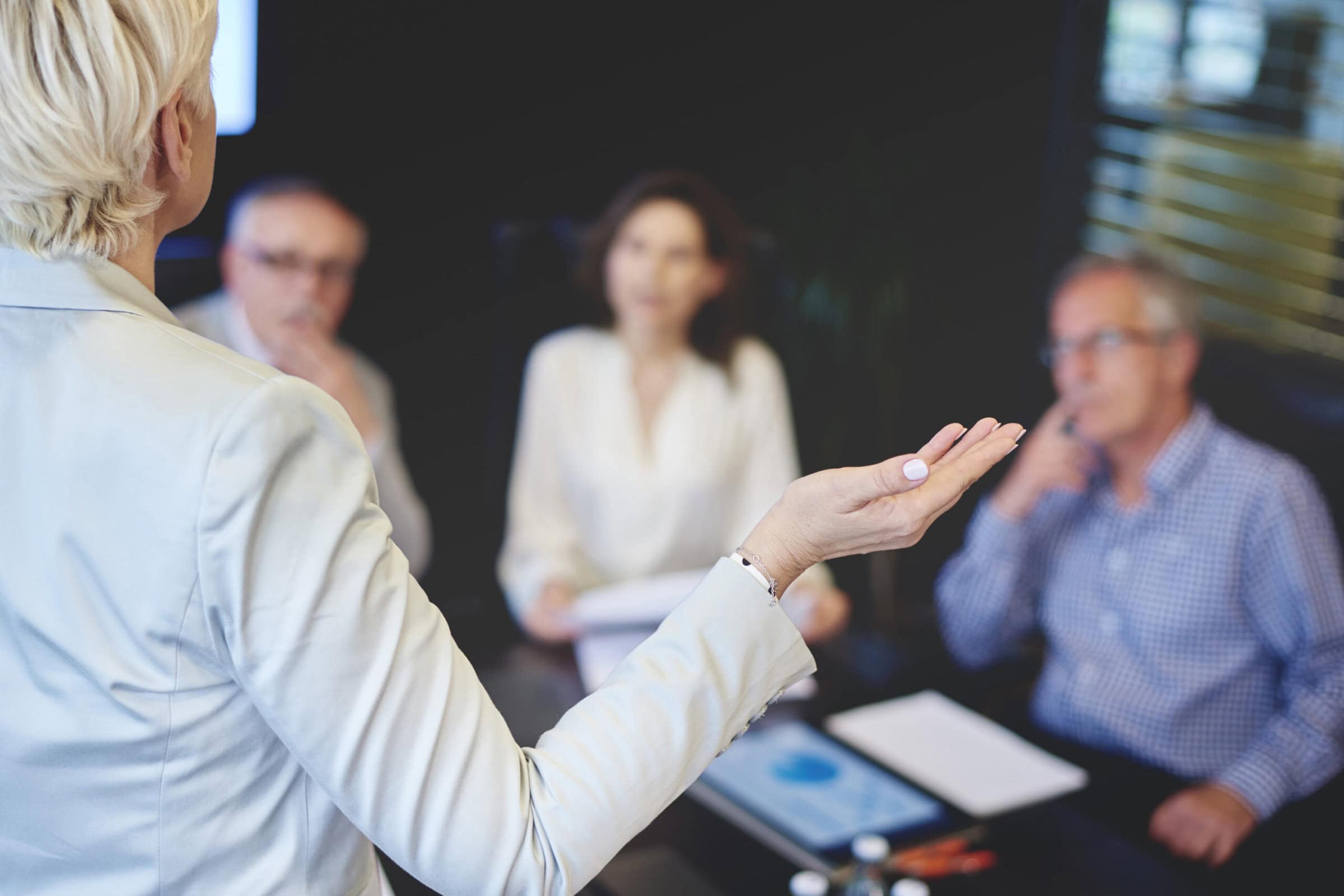 A professional woman in a business suit confidently presents to an audience during a meeting.